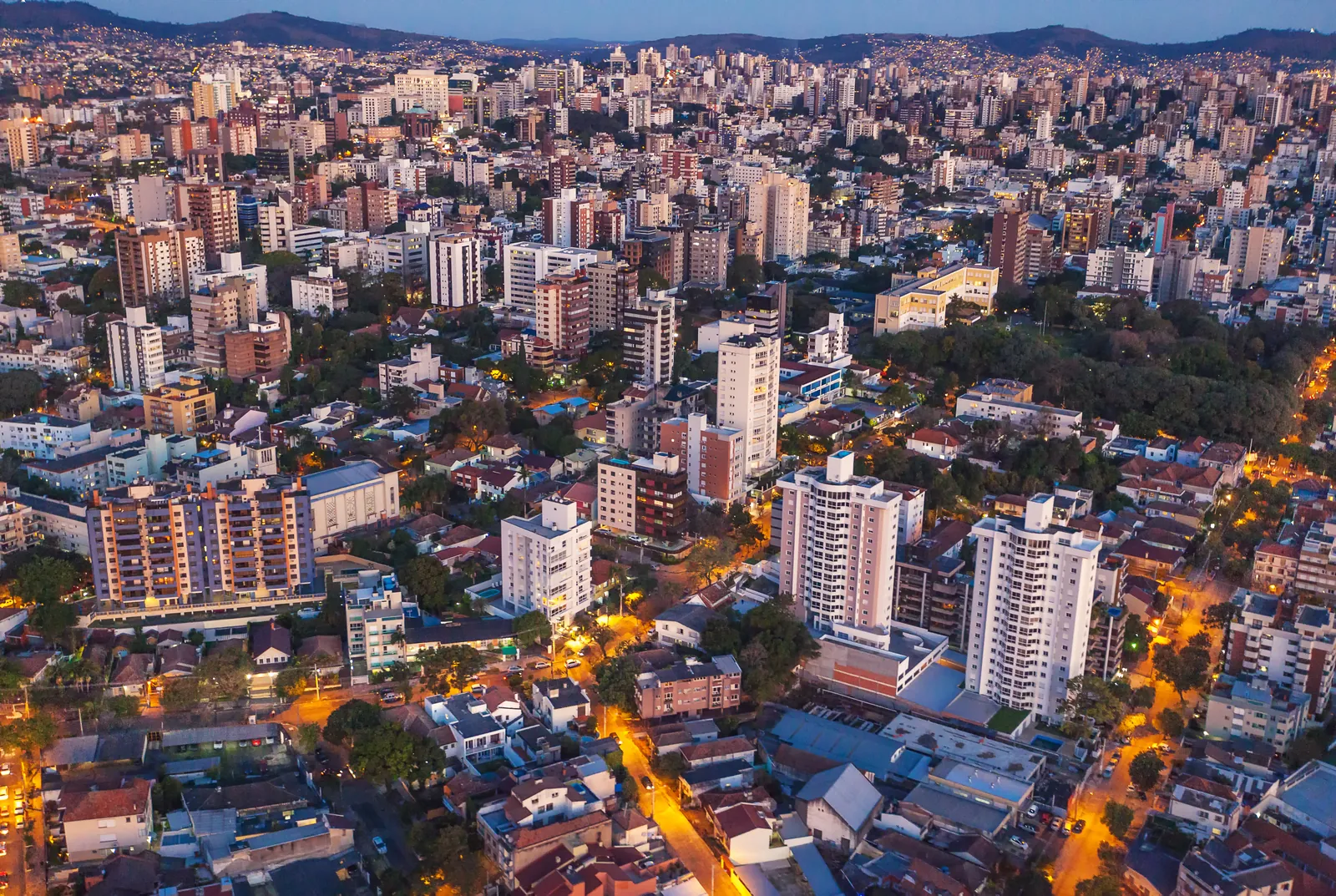 Porto Alegre vista de helicóptero ao anoitecer, com prédios, árvores e ruas iluminadas.