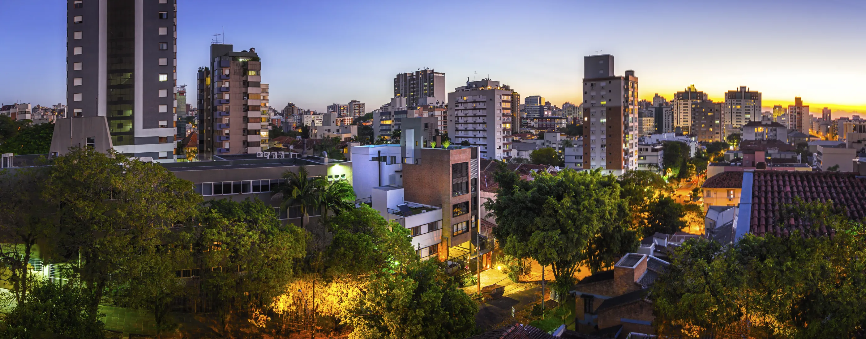 Foto panorâmica de Porto Alegre ao anoitecer, com o Edifício Casa América, de tijolos, entre as árvores e os prédios.