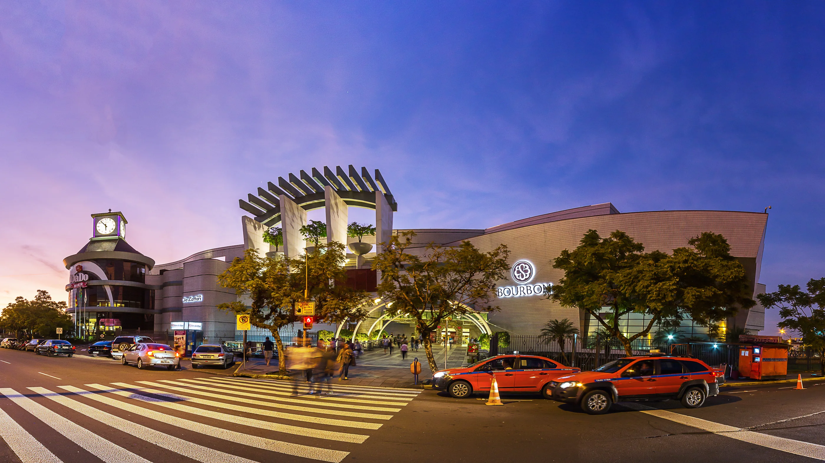Fachada do Bourbon Shopping Country ao entardecer, em panorâmica, com céu roxo e azul, a torre do relógio à esquerda e a pérgola no alto.