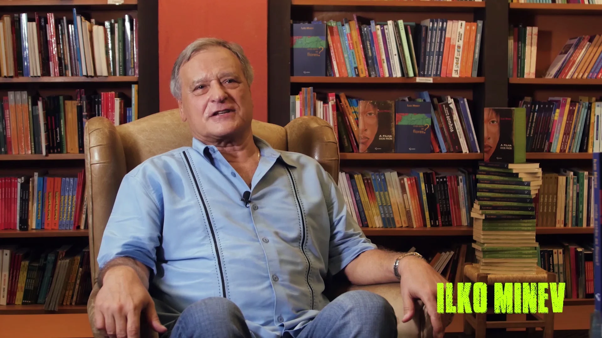 Ilko Minev em entrevista, sentado numa poltrona de couro, diante de estantes cheias de livros, com os livros dele em destaque.