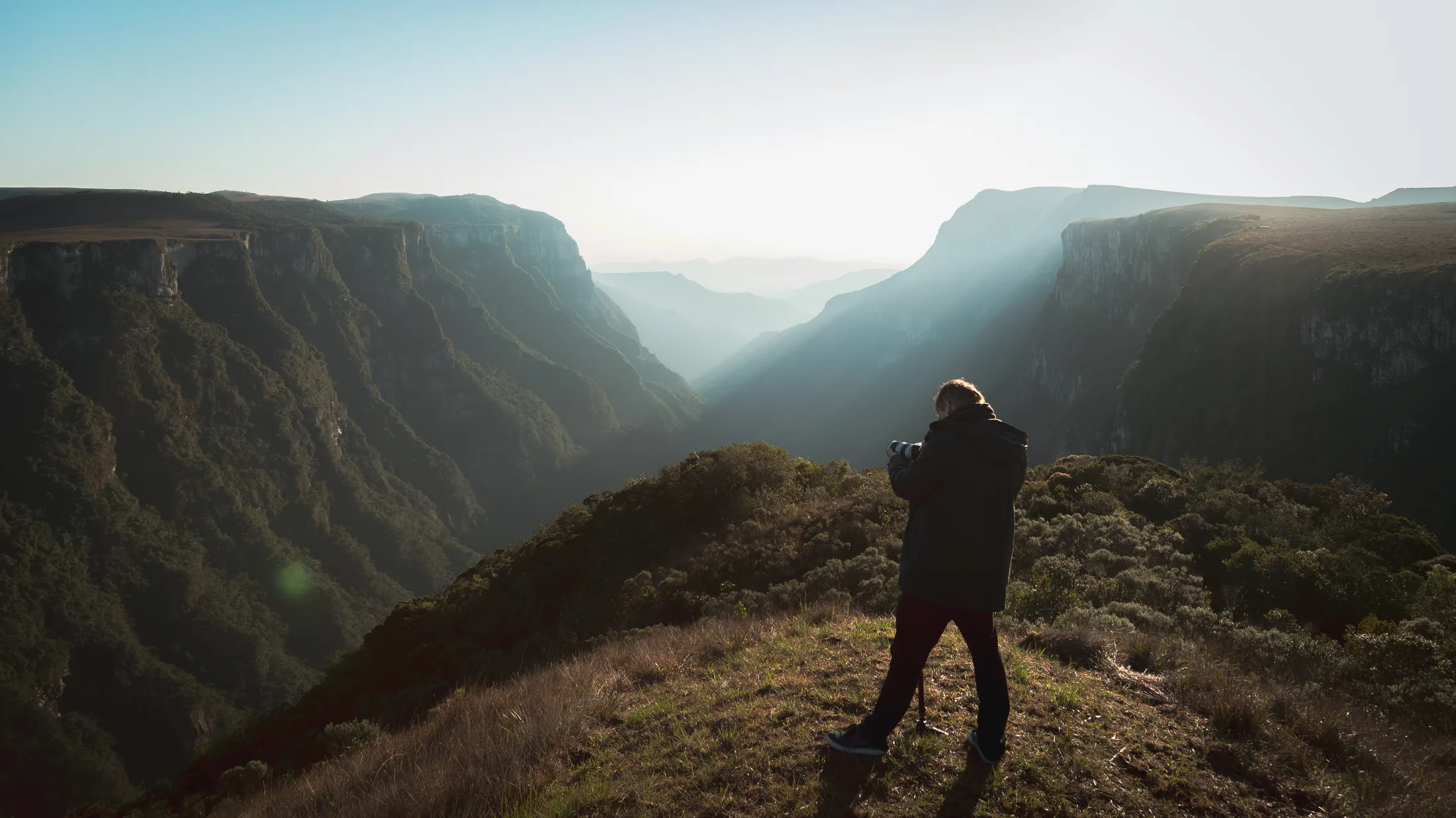 Um fotógrafo de costas, sobre uma elevação, diante de um cânion em Cambará do Sul, com a luz atravessando a neblina.