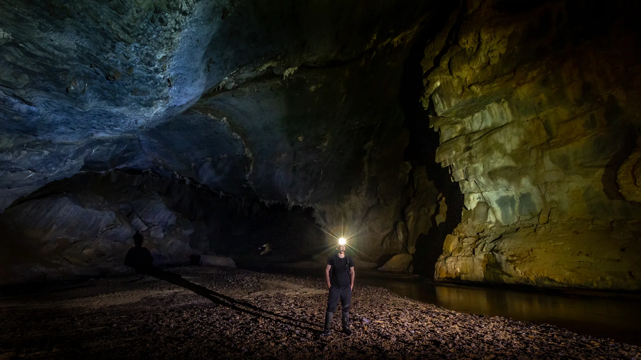 O apresentador com lanterna na testa dentro da Caverna Temimina, diante de uma parede de rocha e de um lago.