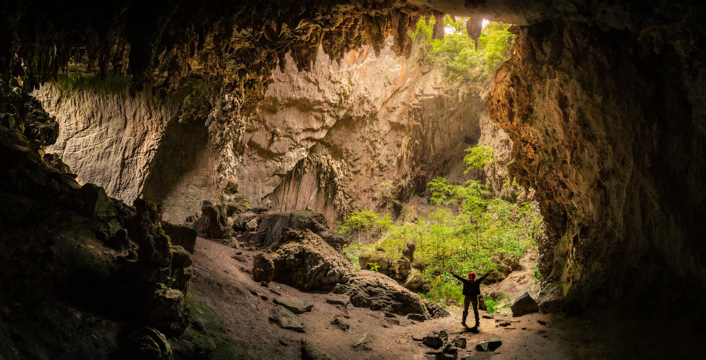 A entrada da Caverna Temimina, com vegetação verde e luz entrando, e uma pessoa de braços abertos ao fundo.