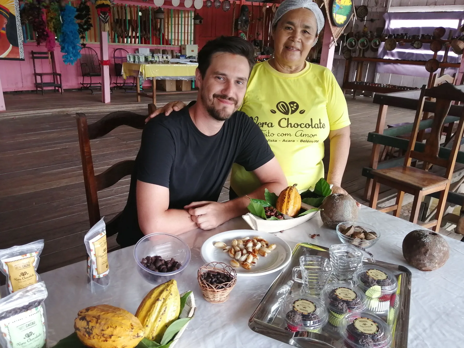 O apresentador ao lado de uma moradora da Ilha Boa Vista do Acará, à mesa com chocolates e cacau.