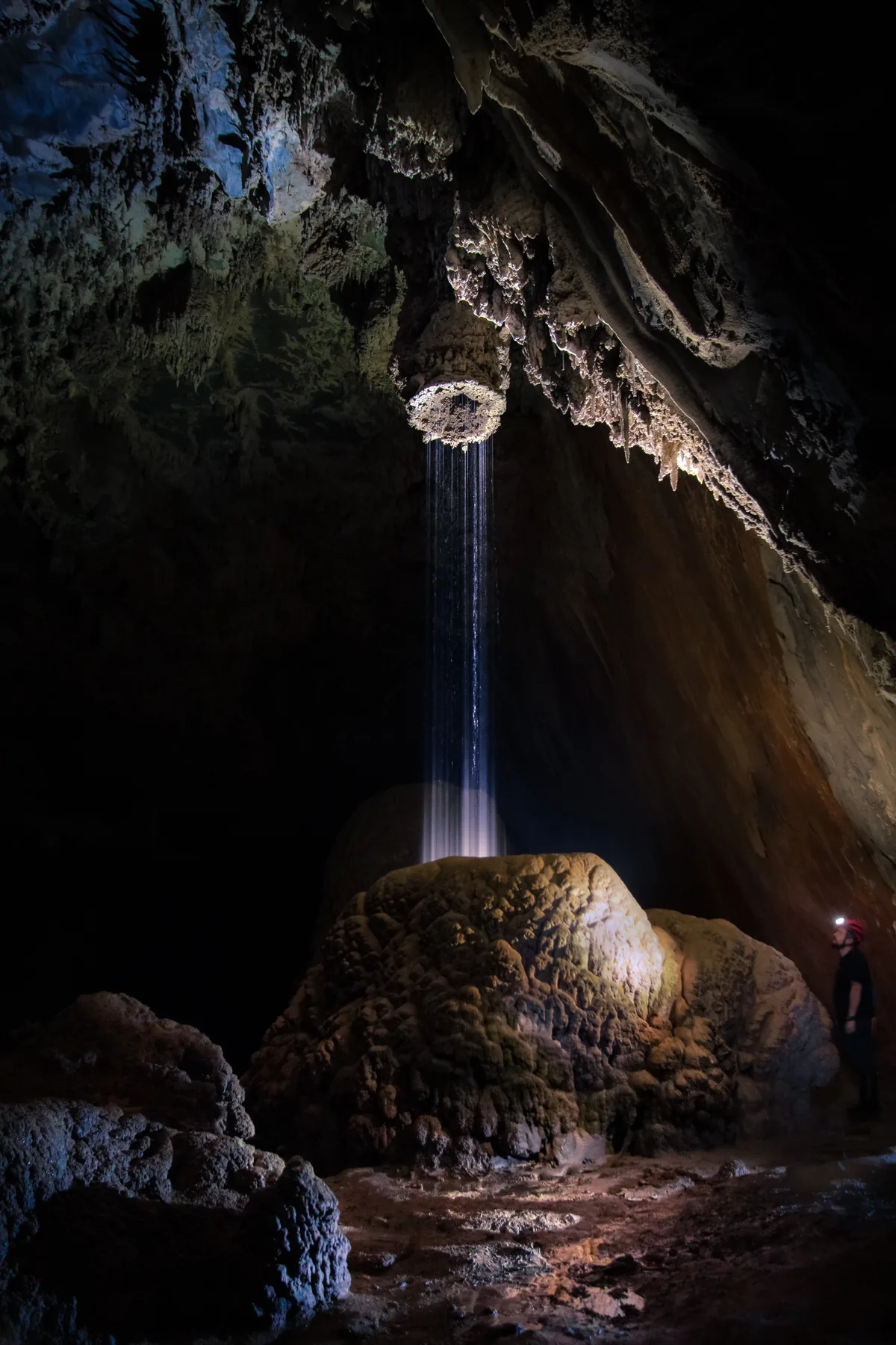 Uma queda d’água dentro de uma caverna em Iporanga, iluminada por um feixe de luz, com uma pessoa à direita.
